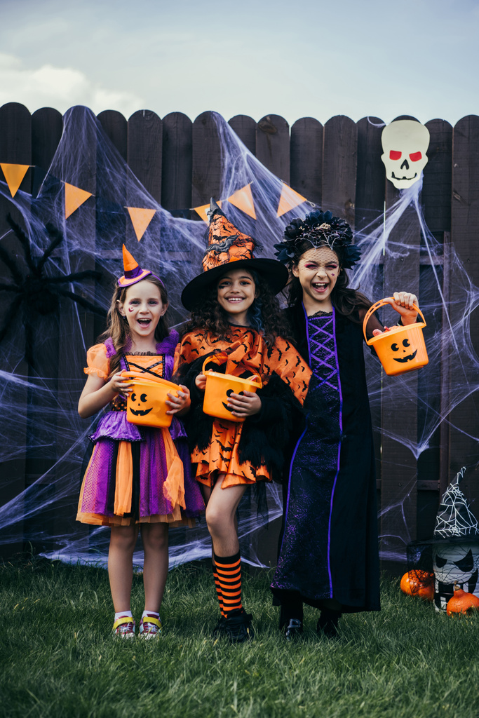 Ragazze sorridenti in costume che tengono fibbie con caramelle vicino decorazione di Halloween e ragnatela sulla recinzione all'aperto - Foto, immagini