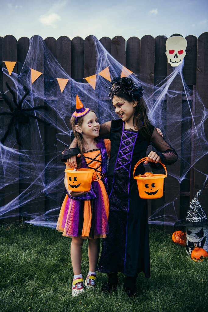 Mosolygó kiskorú gyerekek ölelkeznek és halloween vödröket tartanak a kertben - Fotó, kép