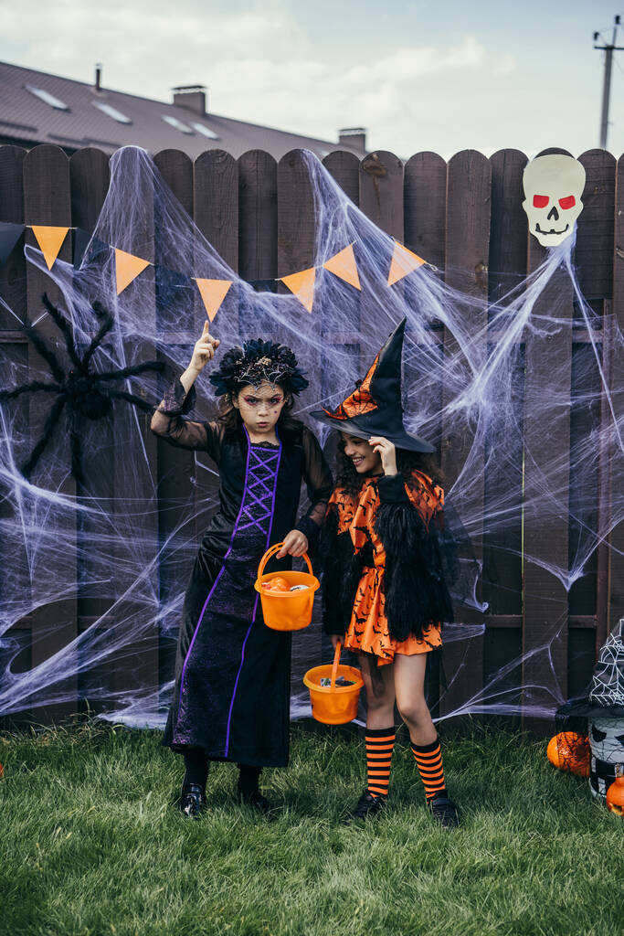 Mädchen im Halloween-Kostüm hält Eimer mit Bonbons neben lächelndem Freund im Hinterhof - Foto, Bild