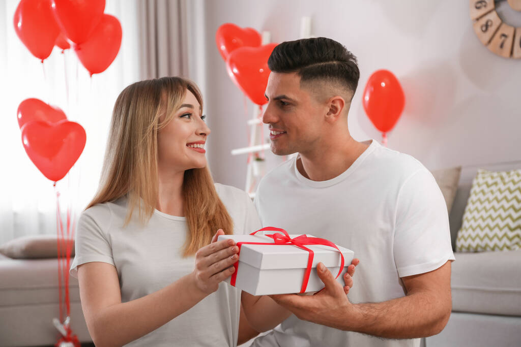 Ο άνθρωπος παρουσιάζει δώρο στην κοπέλα του σε δωμάτιο διακοσμημένο με μπαλόνια σε σχήμα καρδιάς. Γιορτή του Αγίου Βαλεντίνου - Φωτογραφία, εικόνα