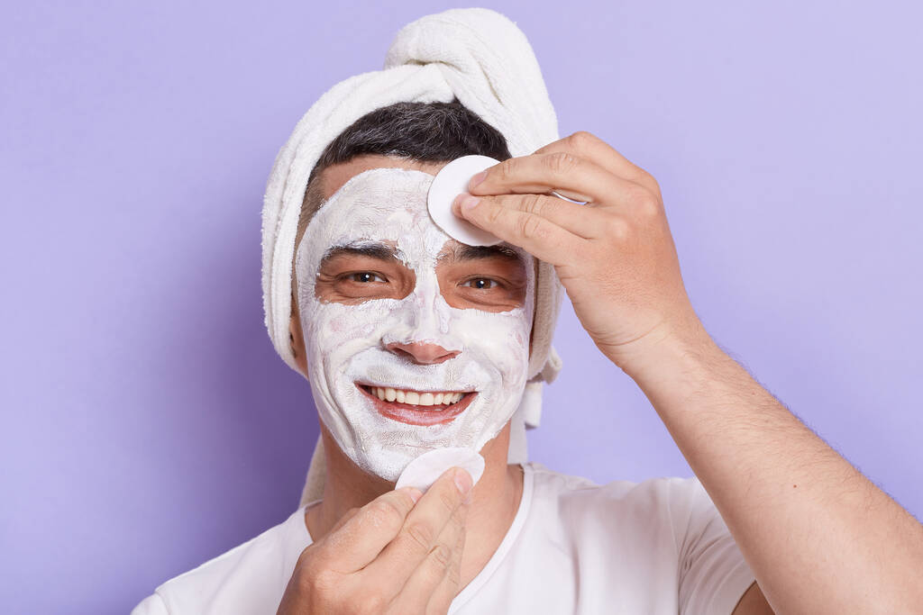 Indoor shot van glimlachende vrolijke blanke man met masker voor de huid doen cosmetologie procedures, verpakt in handdoek op het hoofd, glimlachen naar de camera, geïsoleerd over violette achtergrond. - Foto, afbeelding