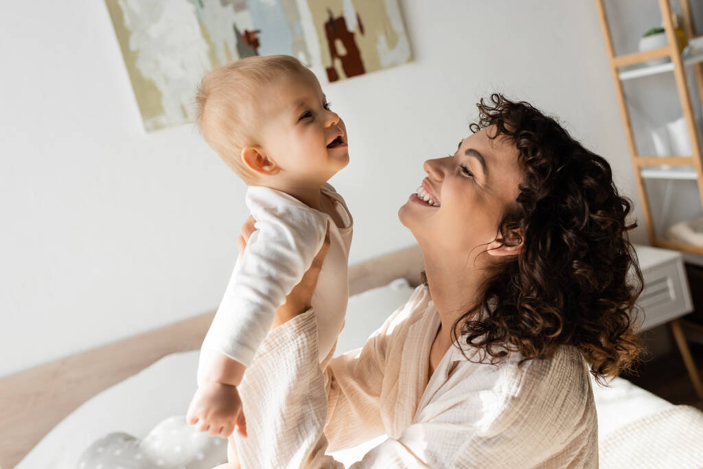 mujer alegre y rizada en ropa de salón sosteniendo en brazos bebé hija en mameluco  - Foto, imagen