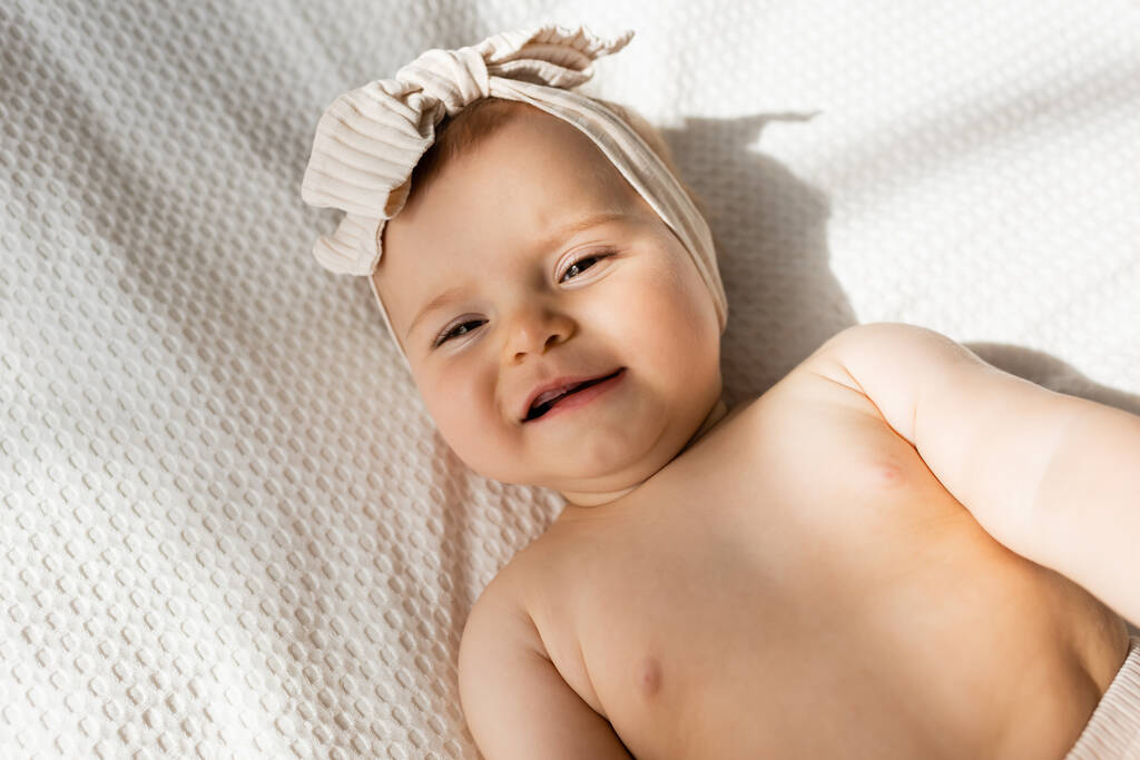 top view of infant girl in headband lying on bed and looking at camera - Photo, Image