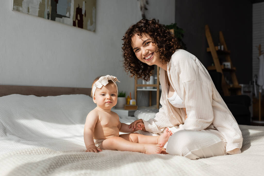 Χαρούμενη μητέρα με σγουρά μαλλιά κάθεται στο κρεβάτι με την κόρη του μωρού στο κεφαλόδεσμο  - Φωτογραφία, εικόνα