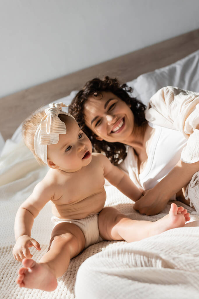 fröhliche Mutter mit lockigem Haar liegt auf dem Bett neben süßer Baby-Tochter im Stirnband  - Foto, Bild