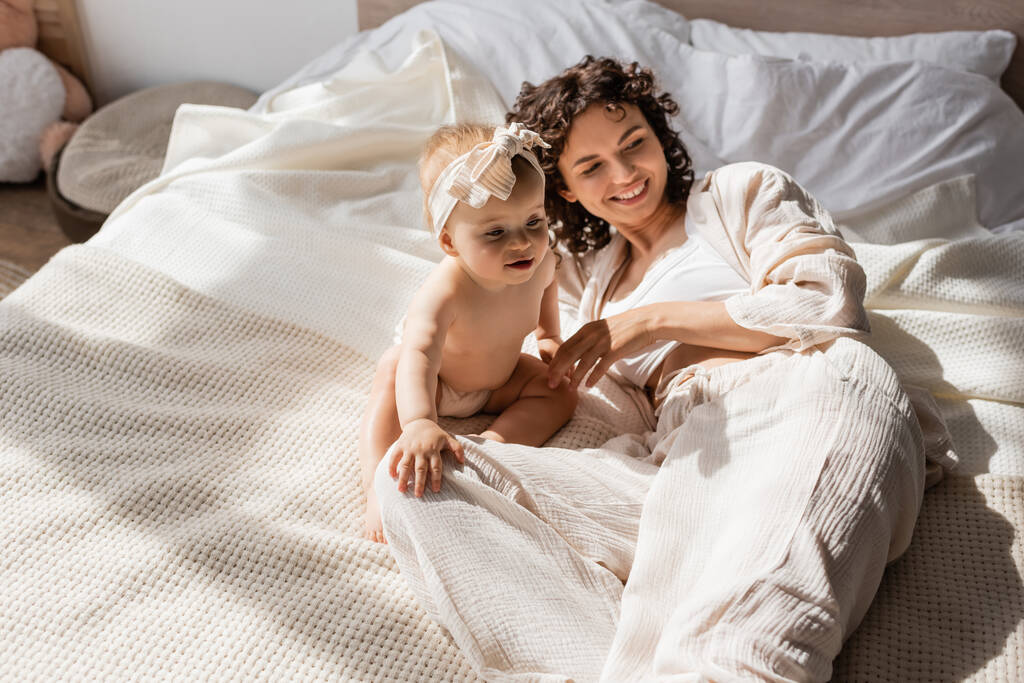 szczęśliwa matka z kręconymi włosami leżące na łóżku w pobliżu słodkie dziecko córka w opasce  - Zdjęcie, obraz