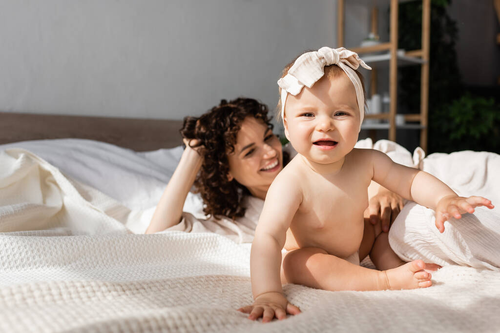 baby meisje in hoofdband zitten op bed in de buurt gelukkig moeder met krullend haar  - Foto, afbeelding
