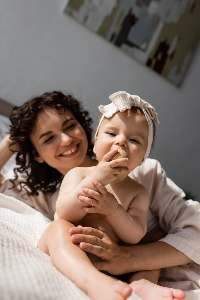 gioiosa donna con i capelli ricci sdraiata sul letto e guardando la figlia neonata in fascia con fiocco - Foto, immagini