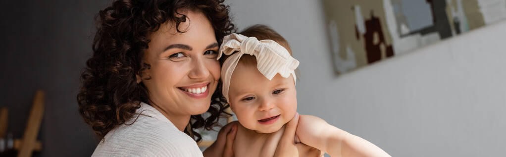 cheerful and curly mother holding in arms baby girl in headband with bow, banner - Photo, Image