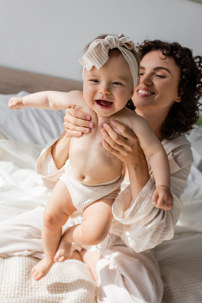 alegre mãe em loungewear sentado na cama e segurando feliz criança filha na cabeça - Foto, Imagem