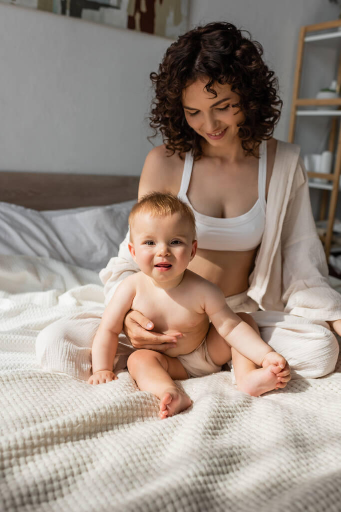 glückliche Mutter in Loungewear mit bauchfreiem Oberteil, die neben ihrer kleinen Tochter im Schlafzimmer sitzt  - Foto, Bild