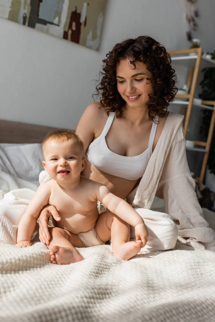 felice madre in loungewear con crop top seduta vicino allegra figlia neonata in camera da letto  - Foto, immagini