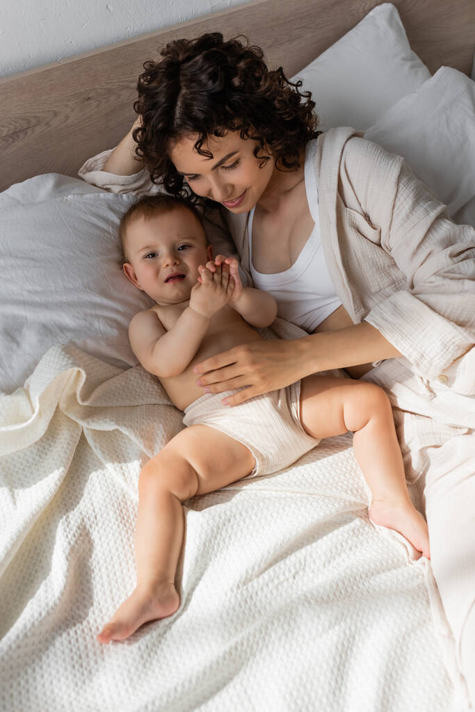 hoge hoek uitzicht van blij krullend moeder in loungewear liggend met baby meisje op bed  - Foto, afbeelding