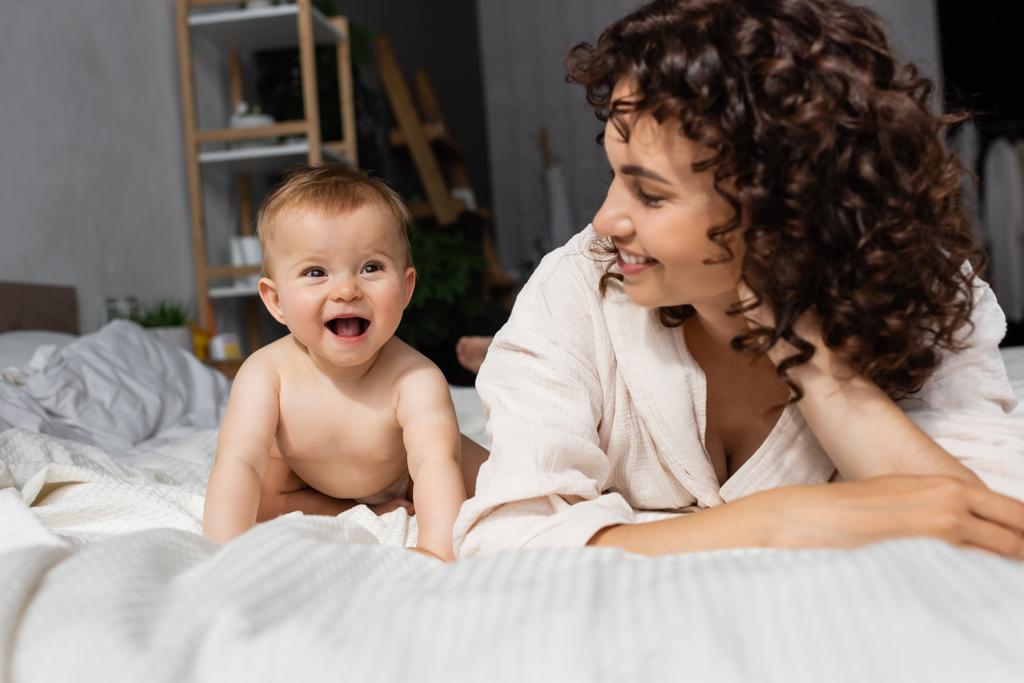 cheerful mother with curly hair looking at cute baby daughter on bed - Photo, Image
