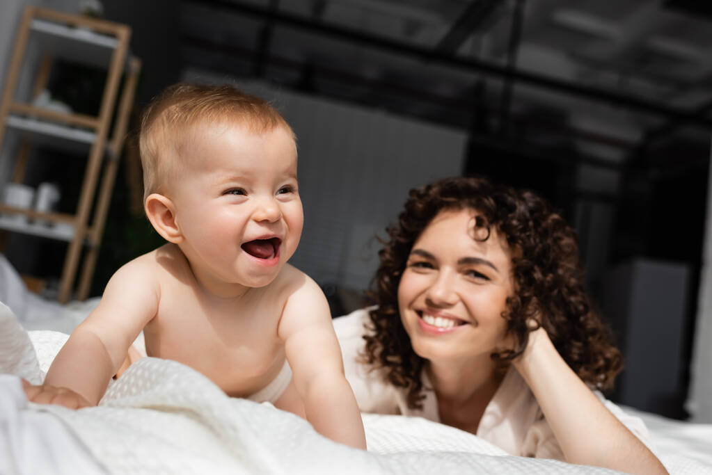 vzrušené dítě dívka plazit na posteli v blízkosti rozmazané matky s kudrnaté vlasy  - Fotografie, Obrázek