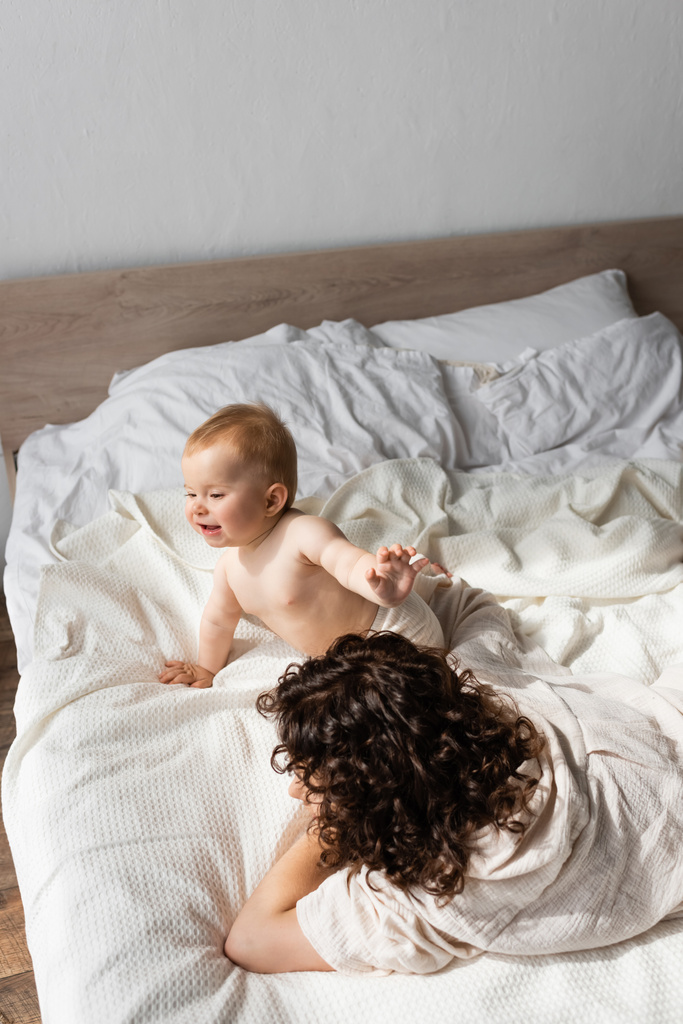 θέα υψηλής γωνίας του κοριτσιού ευτυχής βρέφος σέρνεται κοντά στη μητέρα με σγουρά μαλλιά ξαπλωμένη στο κρεβάτι  - Φωτογραφία, εικόνα