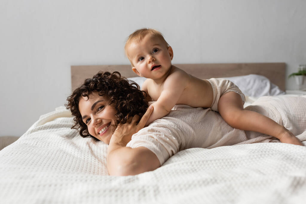 Bebek, yatak odasında kıvırcık saçlı, mutlu bir annenin sırtında yatıyor.  - Fotoğraf, Görsel