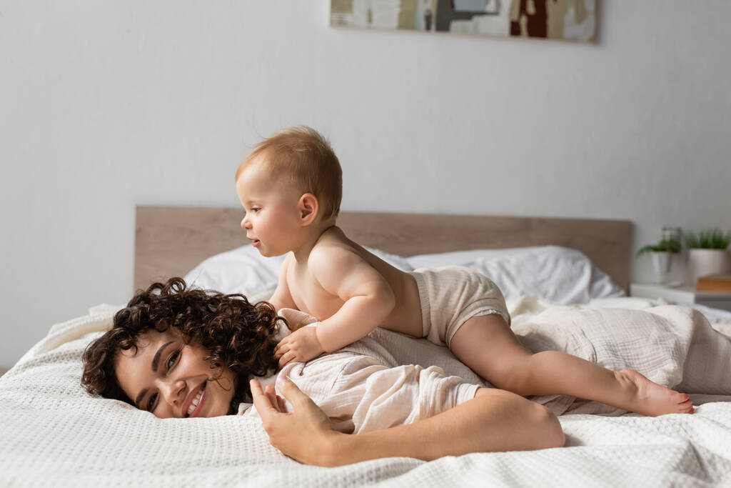 ベッドルームで笑っている母親の後ろに寝そべっている元気な赤ちゃん  - 写真・画像