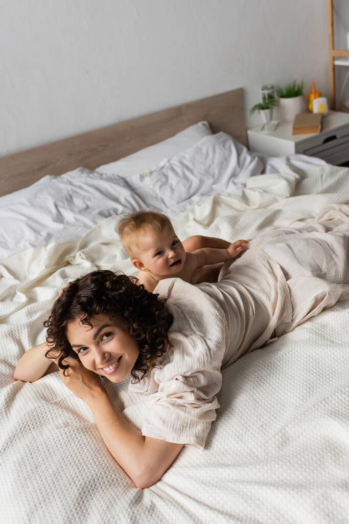 Bebek için yüksek açı yatak odasında neşeli ve kıvırcık bir anne yatıyor.  - Fotoğraf, Görsel