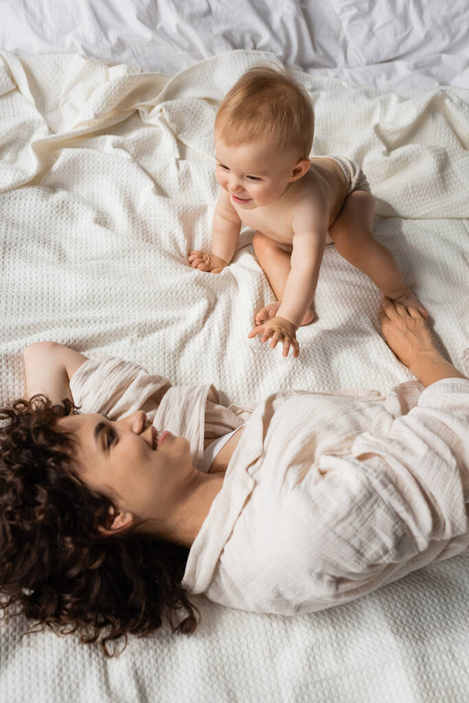 ベッドの上で明るい赤ちゃんの娘を見ながら笑っている母親のトップビュー - 写真・画像