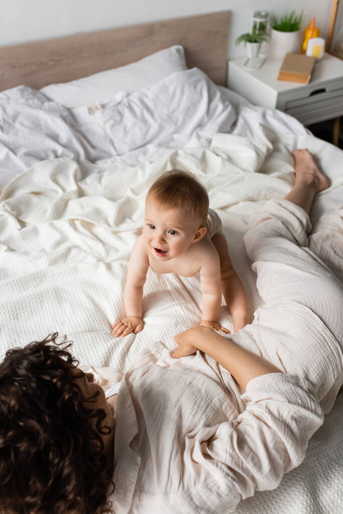 top view of amazed infant kid looking at curly mother resting on bed - Photo, Image