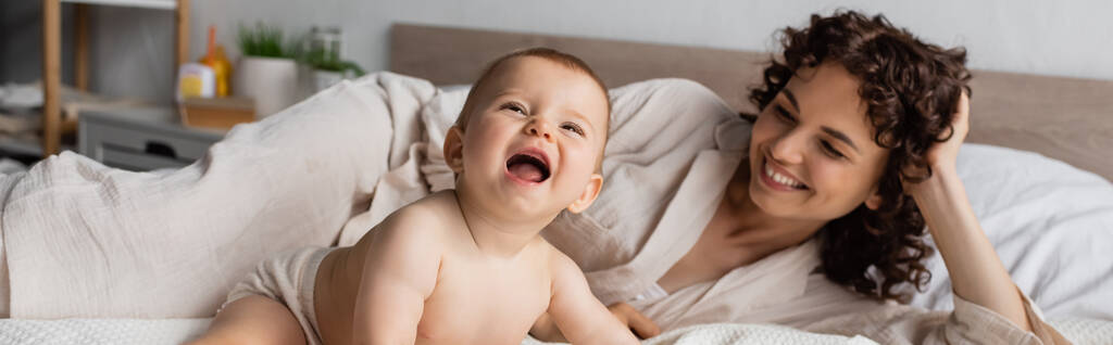glückliche und lockige Mutter lächelt, während sie aufgeregte kleine Tochter auf dem Bett ansieht, Banner - Foto, Bild