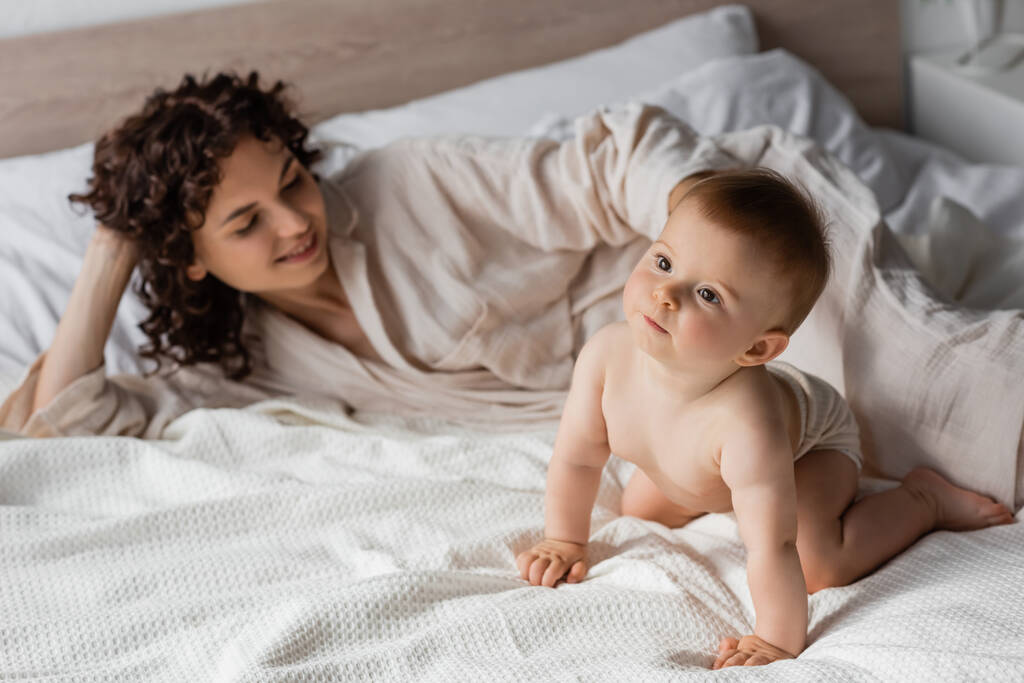 femme positive avec les cheveux bouclés souriant tout en regardant bébé rampant sur le lit - Photo, image