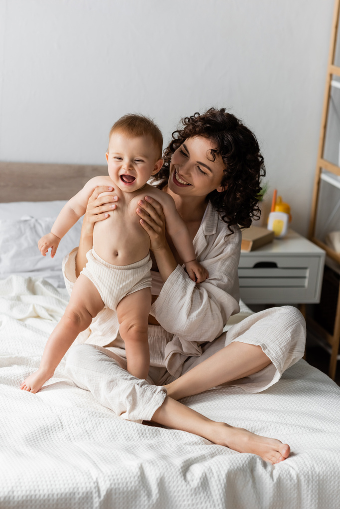 boldog nő göndör haj mosolyog, miközben tartja a karját izgatott csecsemő baba  - Fotó, kép