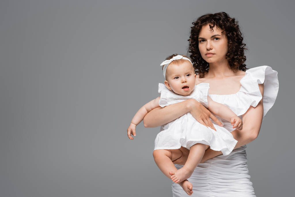 Femme bouclée en robe blanche tenant bébé fille isolé sur gris  - Photo, image