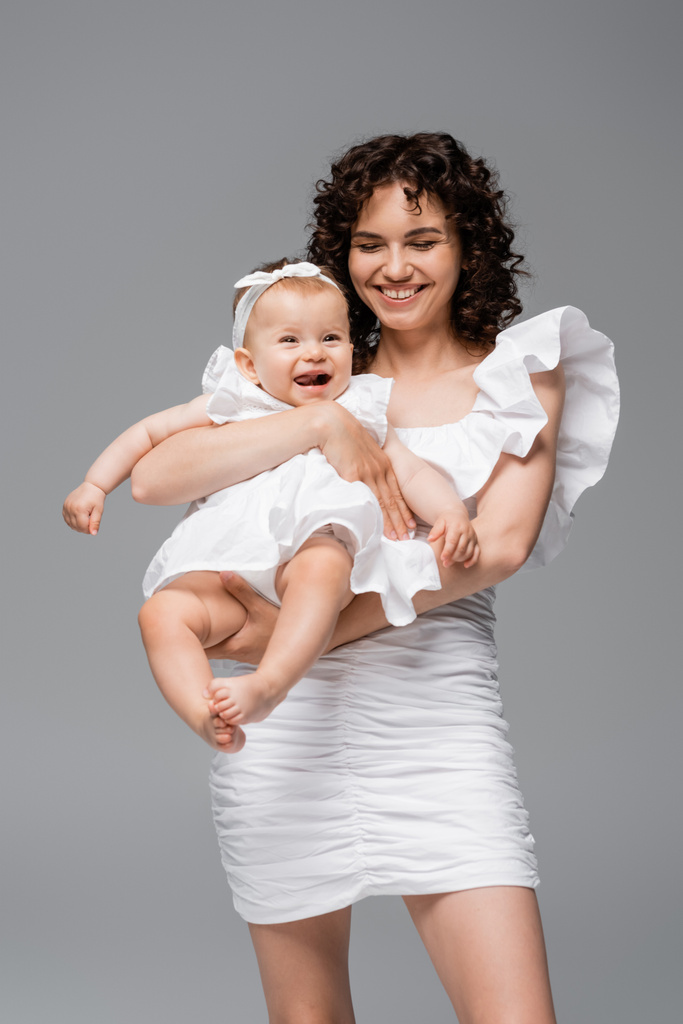 Morena mujer en vestido sosteniendo alegre bebé hija aislado en gris  - Foto, imagen