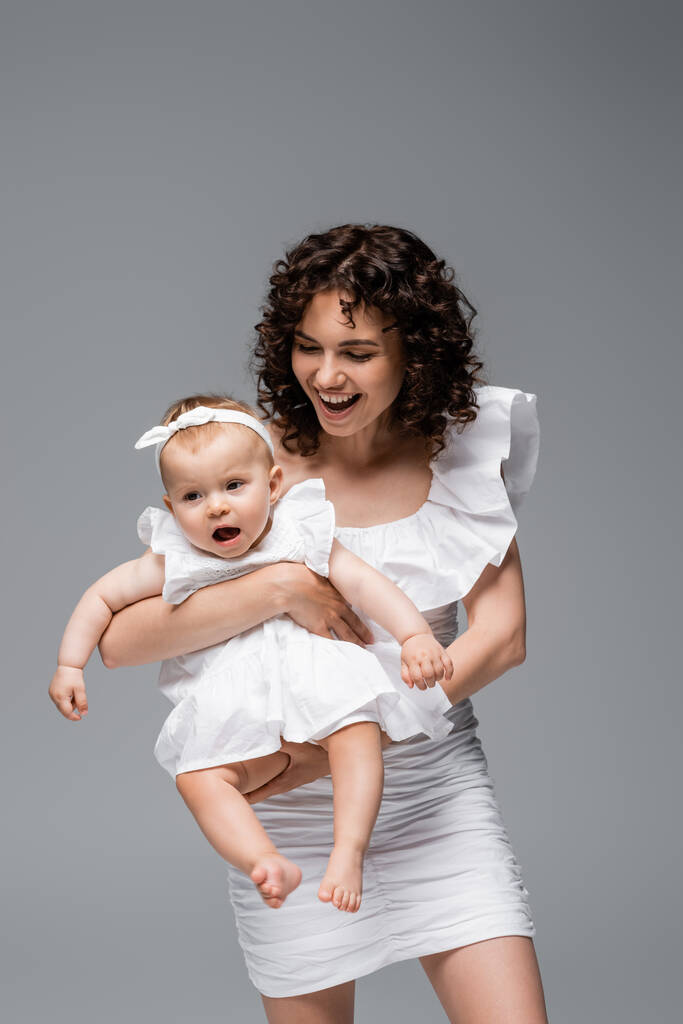Позитивная кудрявая мама держит маленькую дочь в платье, изолированном от серых  - Фото, изображение