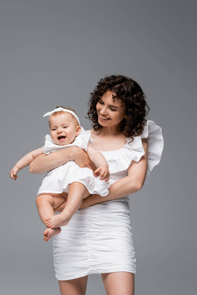 Улыбающаяся кудрявая мать, держа веселого ребенка в платье, изолированном от серых  - Фото, изображение