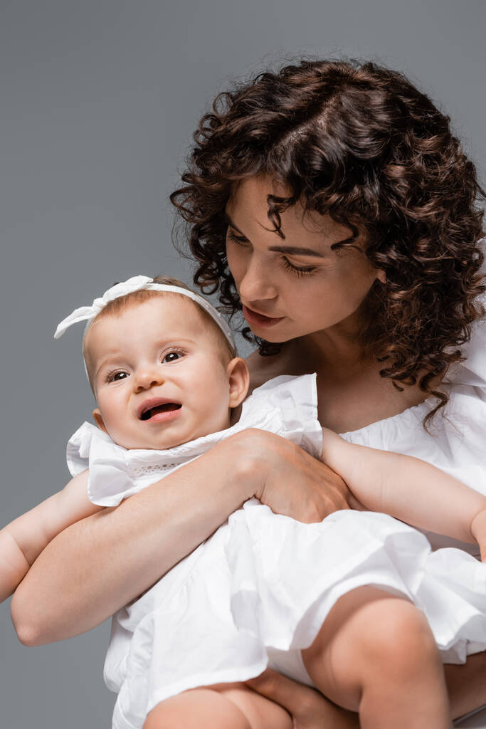 krullend ouder kijken naar baby meisje in wit hoofdband en jurk geïsoleerd op grijs  - Foto, afbeelding