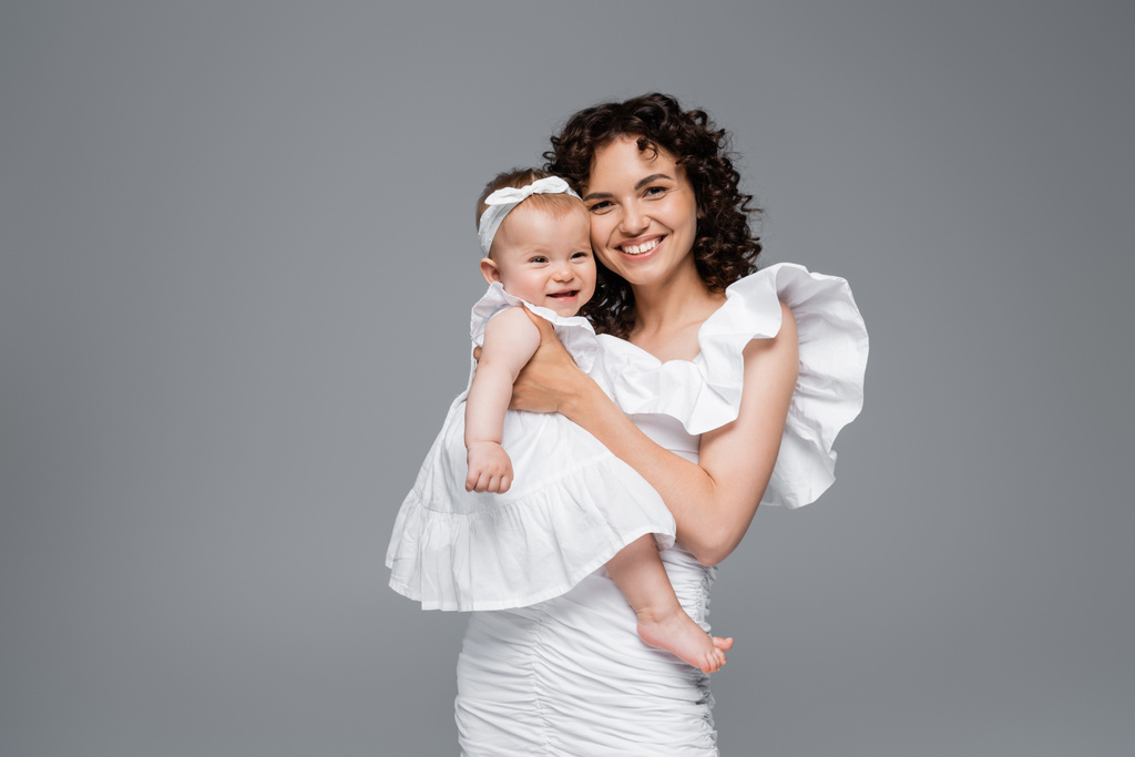 Θετική μητέρα αγκαλιάζει χαρούμενο βρέφος κόρη σε φόρεμα απομονωμένο σε γκρι  - Φωτογραφία, εικόνα