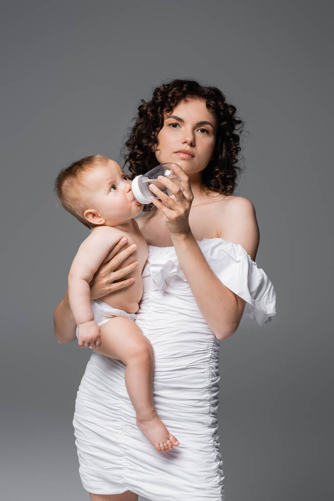 Trendy vrouw in witte jurk houden dochter en baby fles terwijl kijken naar camera geïsoleerd op grijs  - Foto, afbeelding
