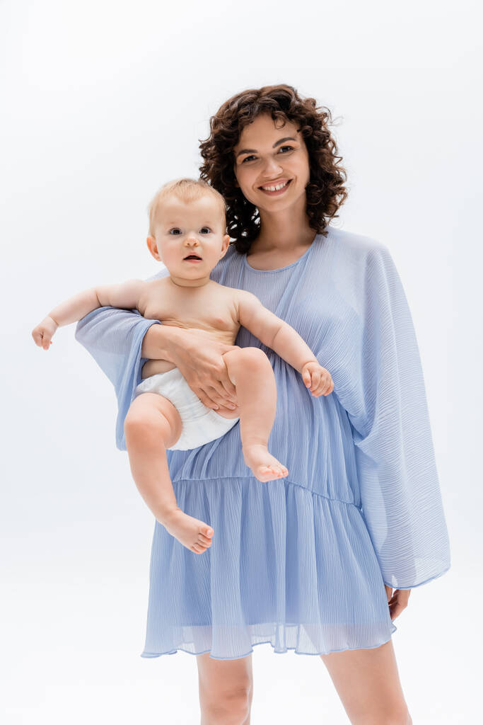 Mulher alegre no vestido segurando bebê e olhando para a câmera isolada no branco  - Foto, Imagem