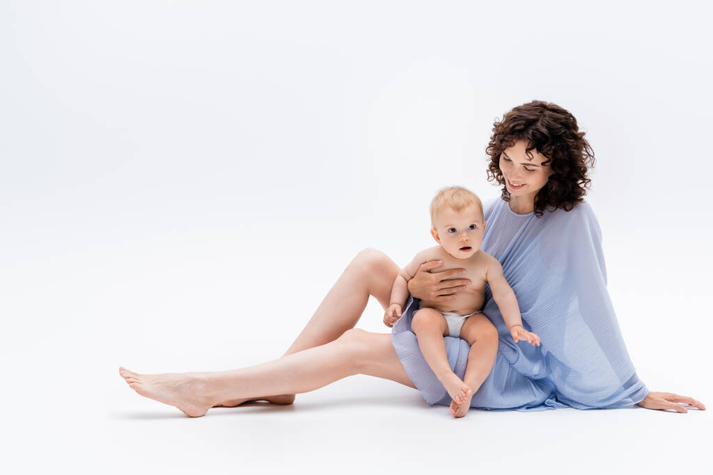 Pleasant mam in blauw jurk aanraken baby meisje terwijl zitten op witte achtergrond  - Foto, afbeelding