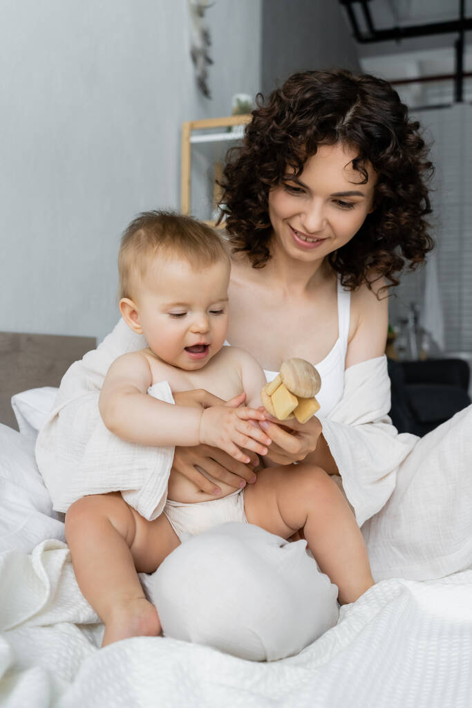 Кучерявая мать держит игрушку рядом с младенческой дочерью на кровати  - Фото, изображение
