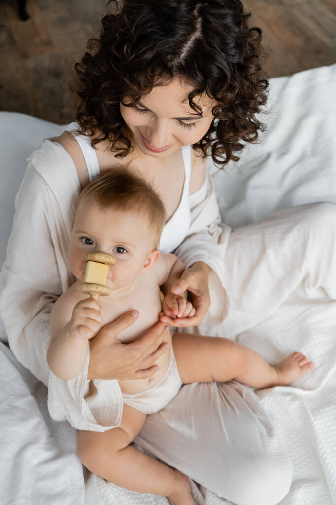 Ansicht einer Frau im Schlafanzug, die ihre kleine Tochter mit Spielzeug auf dem Bett hält  - Foto, Bild