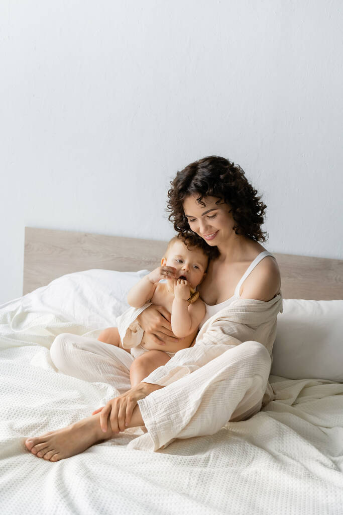 Улыбающаяся женщина обнимает дочку с деревянной игрушкой на кровати  - Фото, изображение