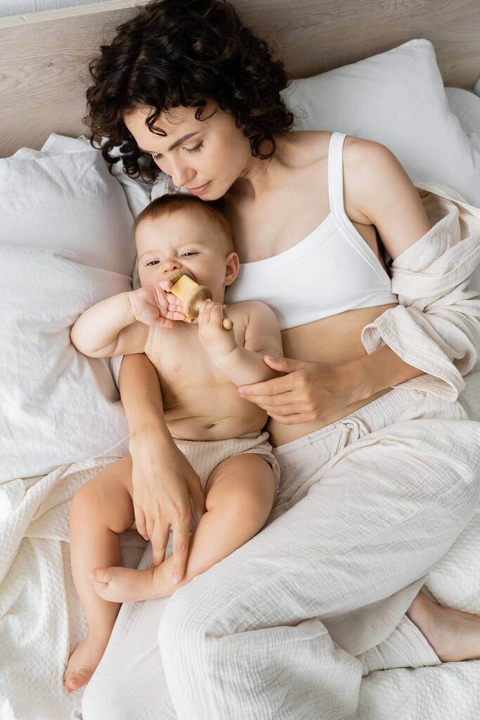 Horní pohled na ženu při pohledu na kojeneckou dívku hrající si s dřevěnou hračkou na posteli  - Fotografie, Obrázek