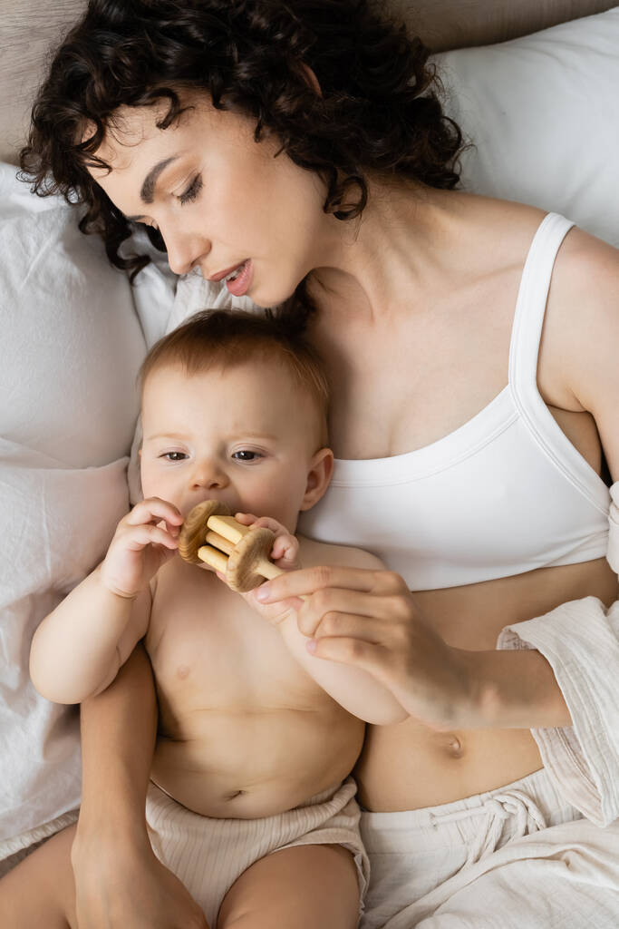 Вид сверху матери в пижаме, держащей игрушку рядом с младенческой дочерью на кровати  - Фото, изображение