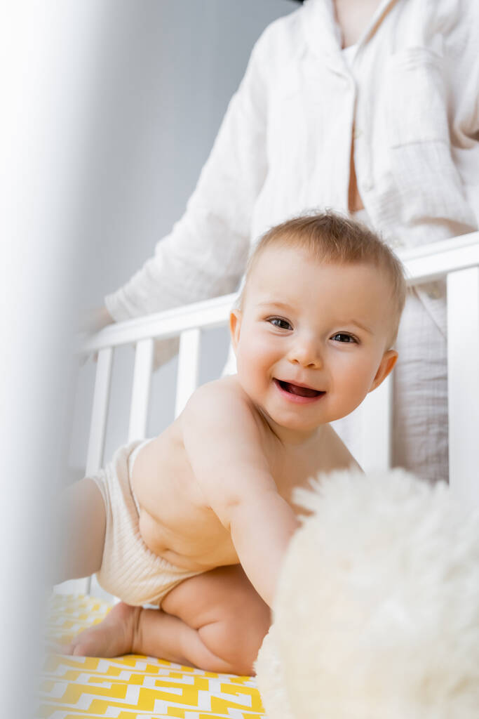 Happy baby girl w majtki patrząc na aparat w pobliżu rozmyte miękkie zabawki w łóżeczko i mama w domu  - Zdjęcie, obraz