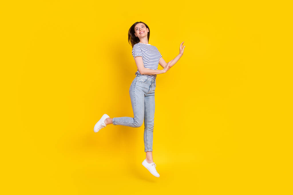 Täysikokoinen kuva ihana nuori nainen näyttää tyhjältä hyppäämällä vapaasti pukeutunut trendikäs raidallinen vaatteita eristetty keltainen väri tausta. - Valokuva, kuva