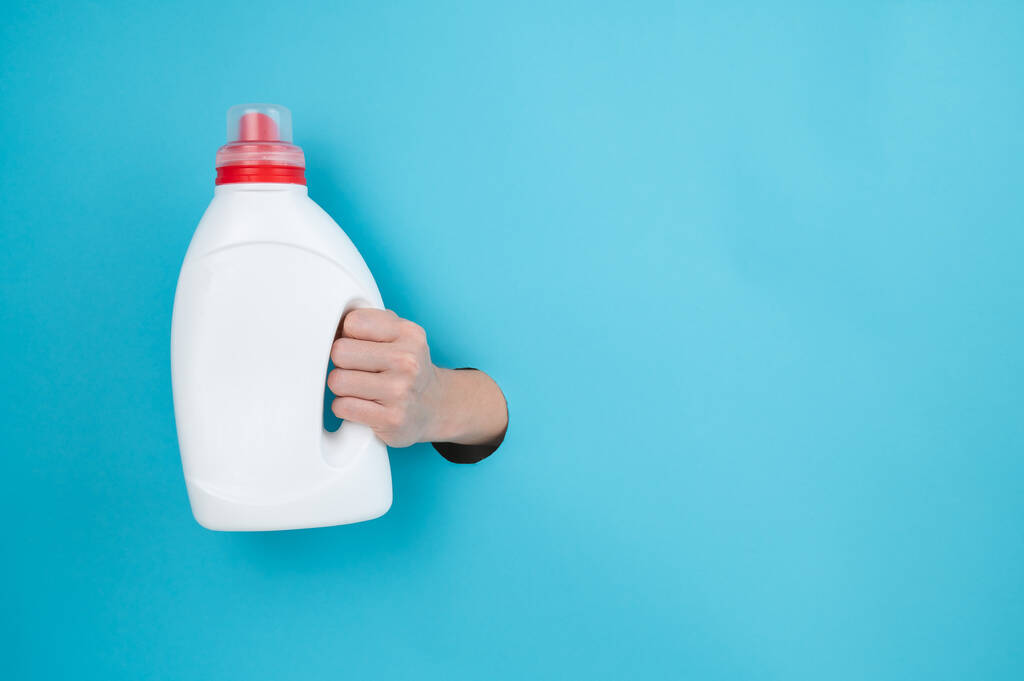 Uma mulher tem uma garrafa branca de detergente. Mão das mulheres saindo do fundo de papel azul - Foto, Imagem