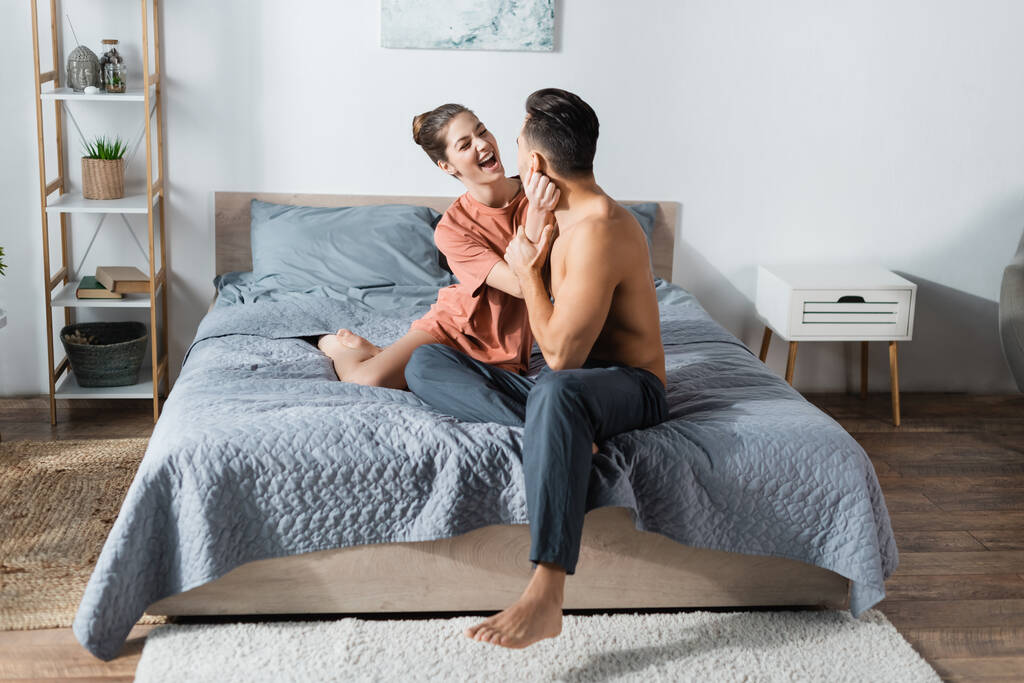 возбужденная женщина в футболке рядом без рубашки мужчина сидит на кровати в пижамных штанах - Фото, изображение