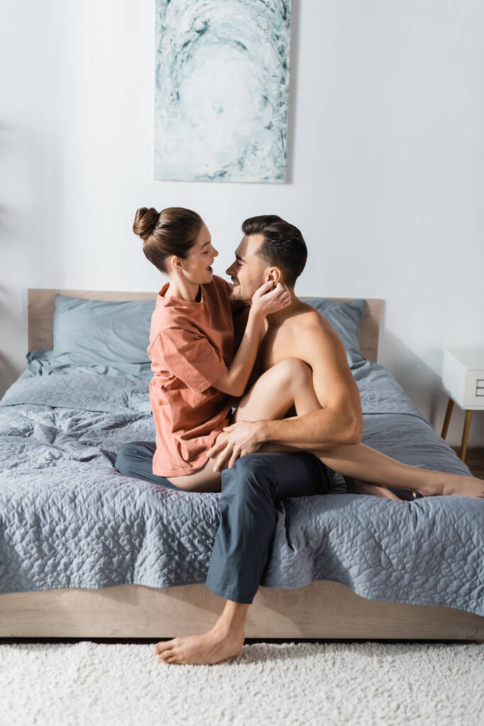 vista laterale della donna in t-shirt che abbraccia con uomo senza camicia in pigiama pantaloni sul letto a casa - Foto, immagini