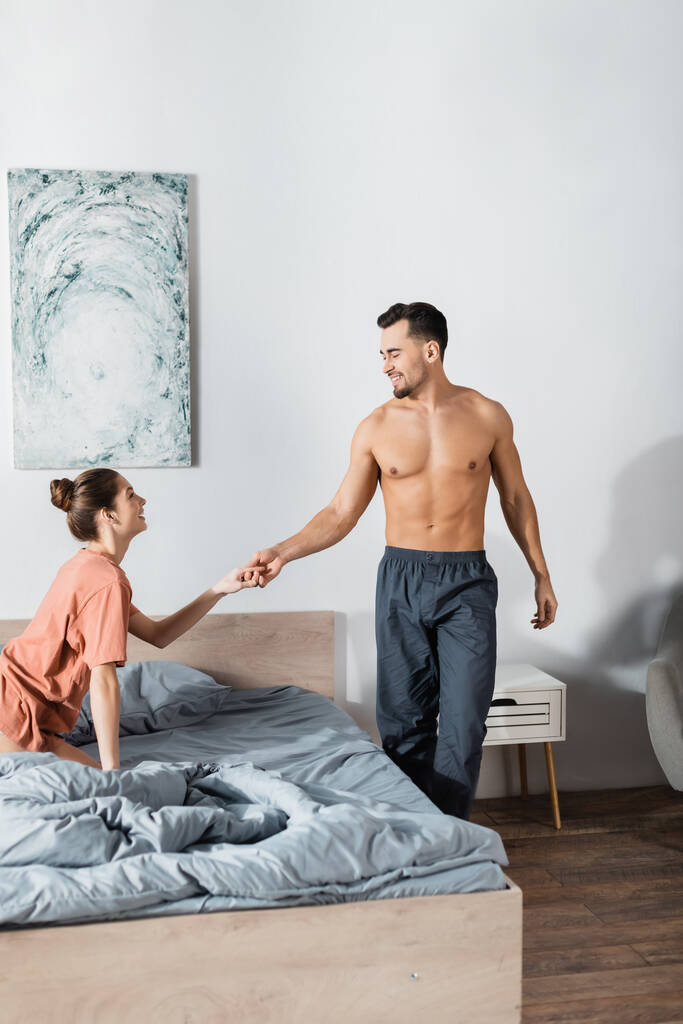 młoda kobieta w t-shircie trzymająca się za ręce z mężczyzną bez koszulki w spodniach piżamy stojącym blisko łóżka - Zdjęcie, obraz