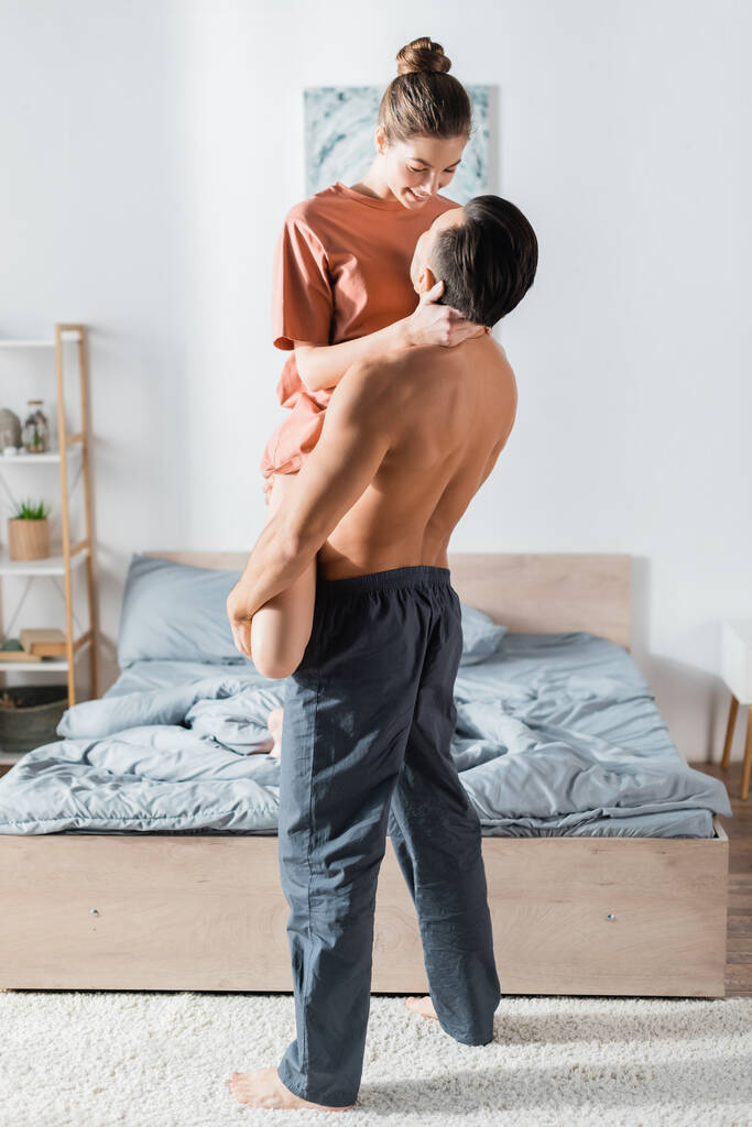 молодой и мускулистый мужчина держит счастливую девушку в руках у кровати дома - Фото, изображение