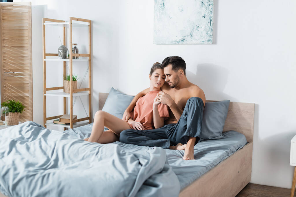 sexy woman in t-shirt holding hands with man in pajama pants while sitting on grey bedding - Photo, Image